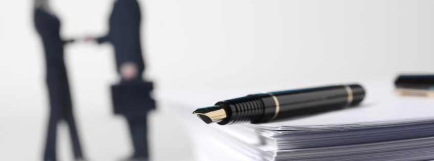 people shaking hands in background, a pen sits on a pile of papers in the foreground