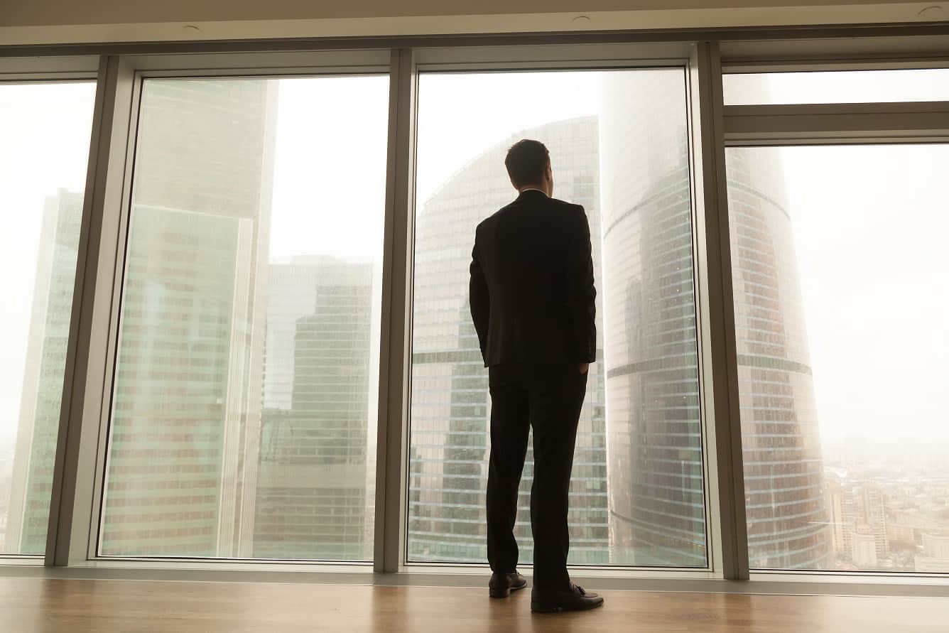 Businessman standing near full-length window looking at city, ba