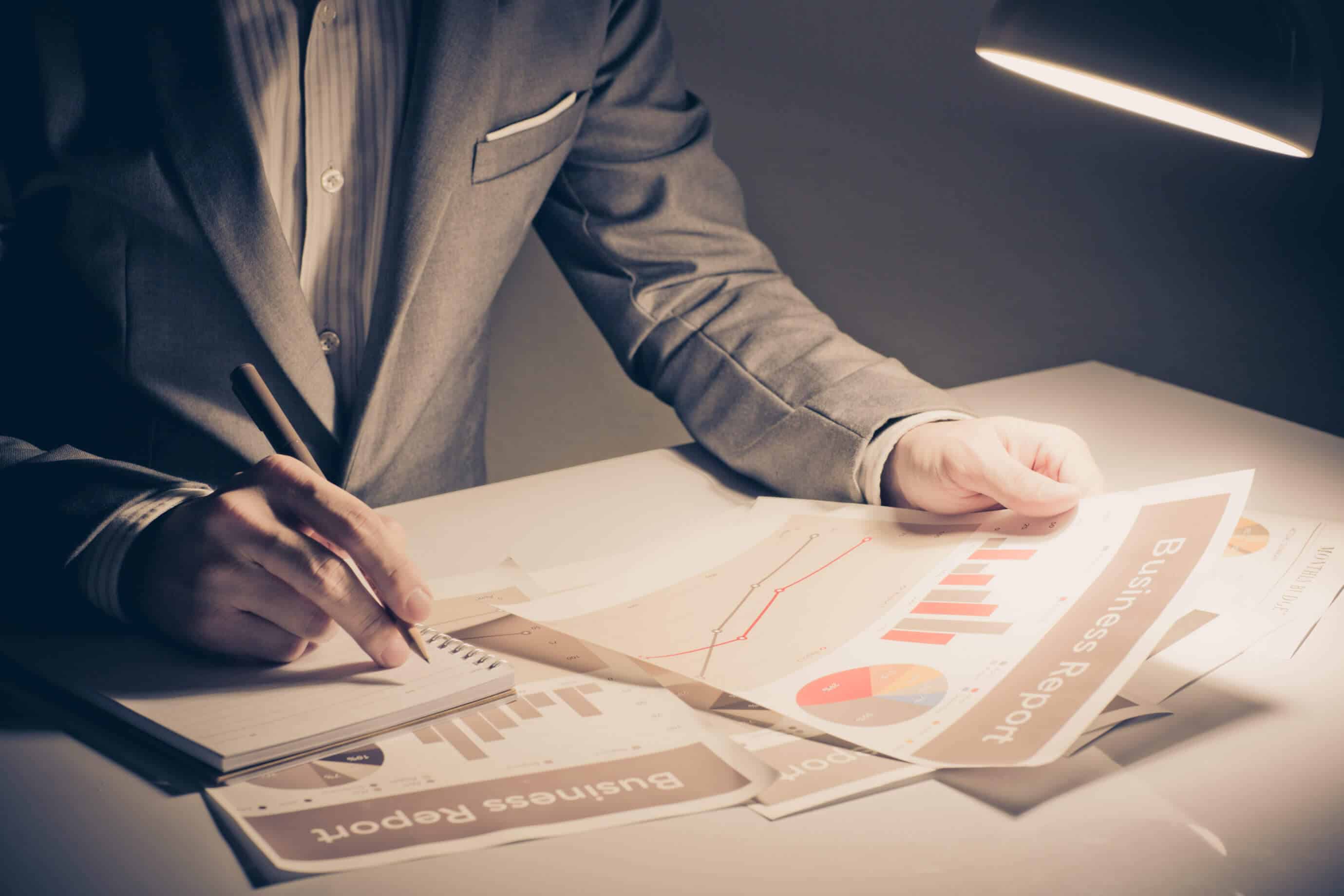 man writing in notepad while reading a business report