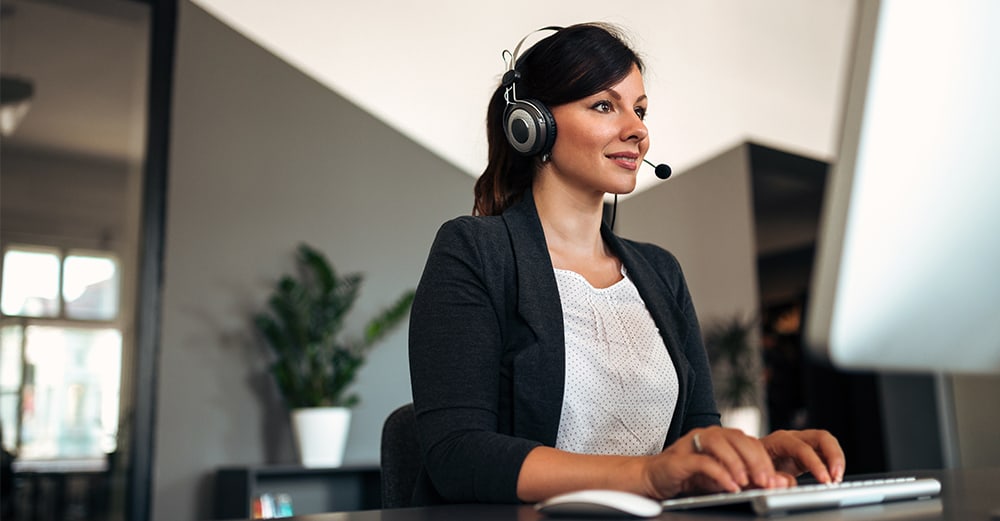 Woman sat at desk in front of a computer wearing a headset as she types on her keyboard