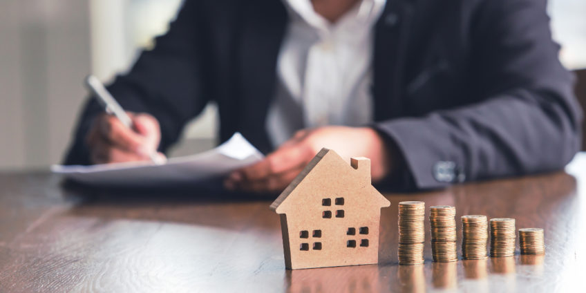 Wooden house with pounds stacked up next to it, and a man signing a contract in the background