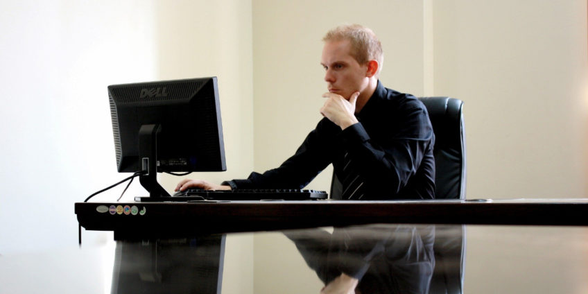 Male business manager wearing suit sat at desk reading on his computer