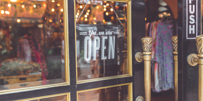 image of a shop window and door with an open sign, inside the shop are clothes hanging up