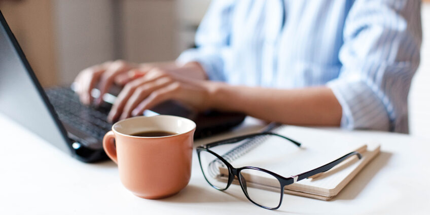 woman working at laptop, with a mug, glasses and notepad resting on the desk beside it